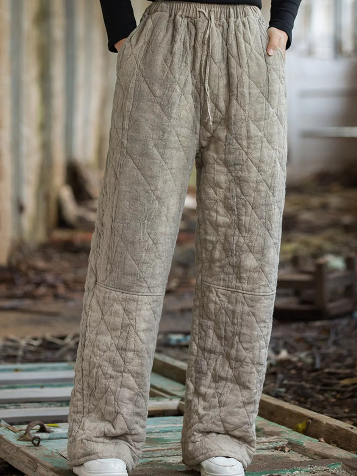 A person wearing loose, quilted wide-leg pants in a light taupe-beige color, standing on a weathered wooden platform in an outdoor rustic setting.
