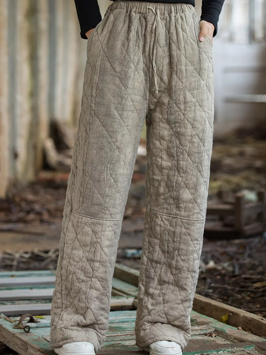 A person wearing loose, quilted wide-leg pants in a light taupe-beige color, standing on a weathered wooden platform in an outdoor rustic setting.