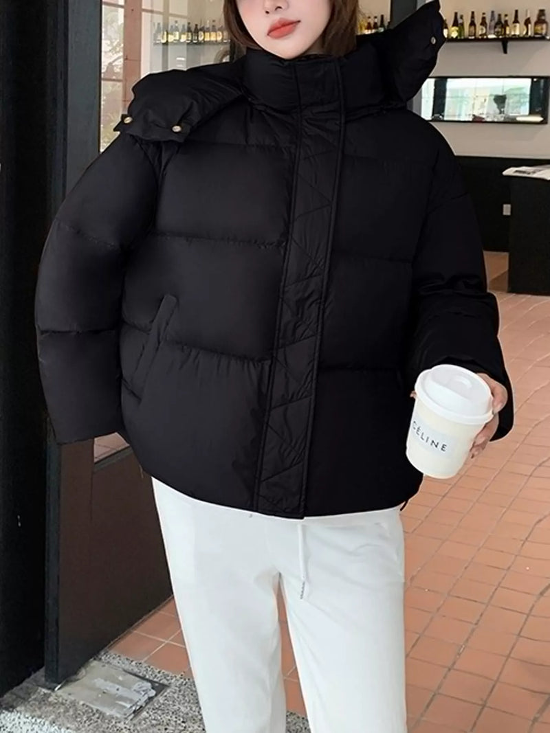 A person wearing a black puffer jacket with a high collar and shoulder detailing, paired with white pants, holding a white coffee cup in a cafe setting.