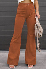 Person wearing brown flared pants holding a beige handbag against a gray wall.