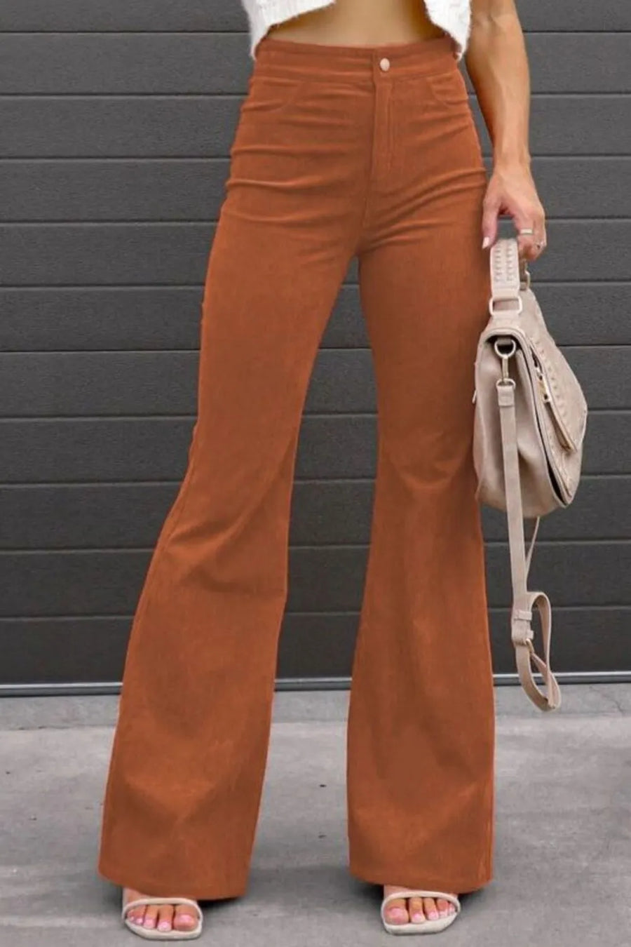 Person wearing brown flared pants holding a beige handbag against a gray wall.