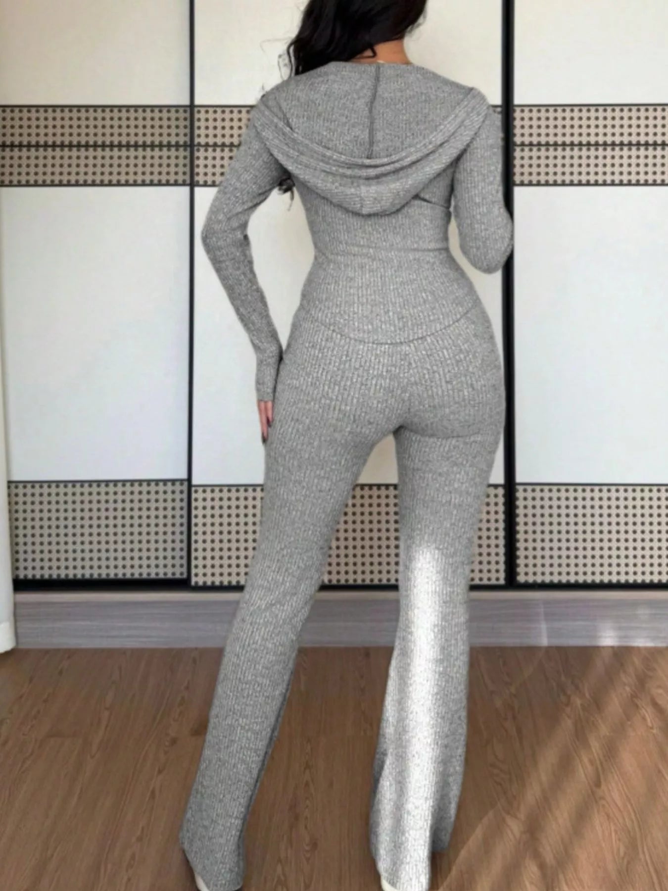 Person wearing a gray knit jumpsuit standing against a white wall with a geometric pattern.