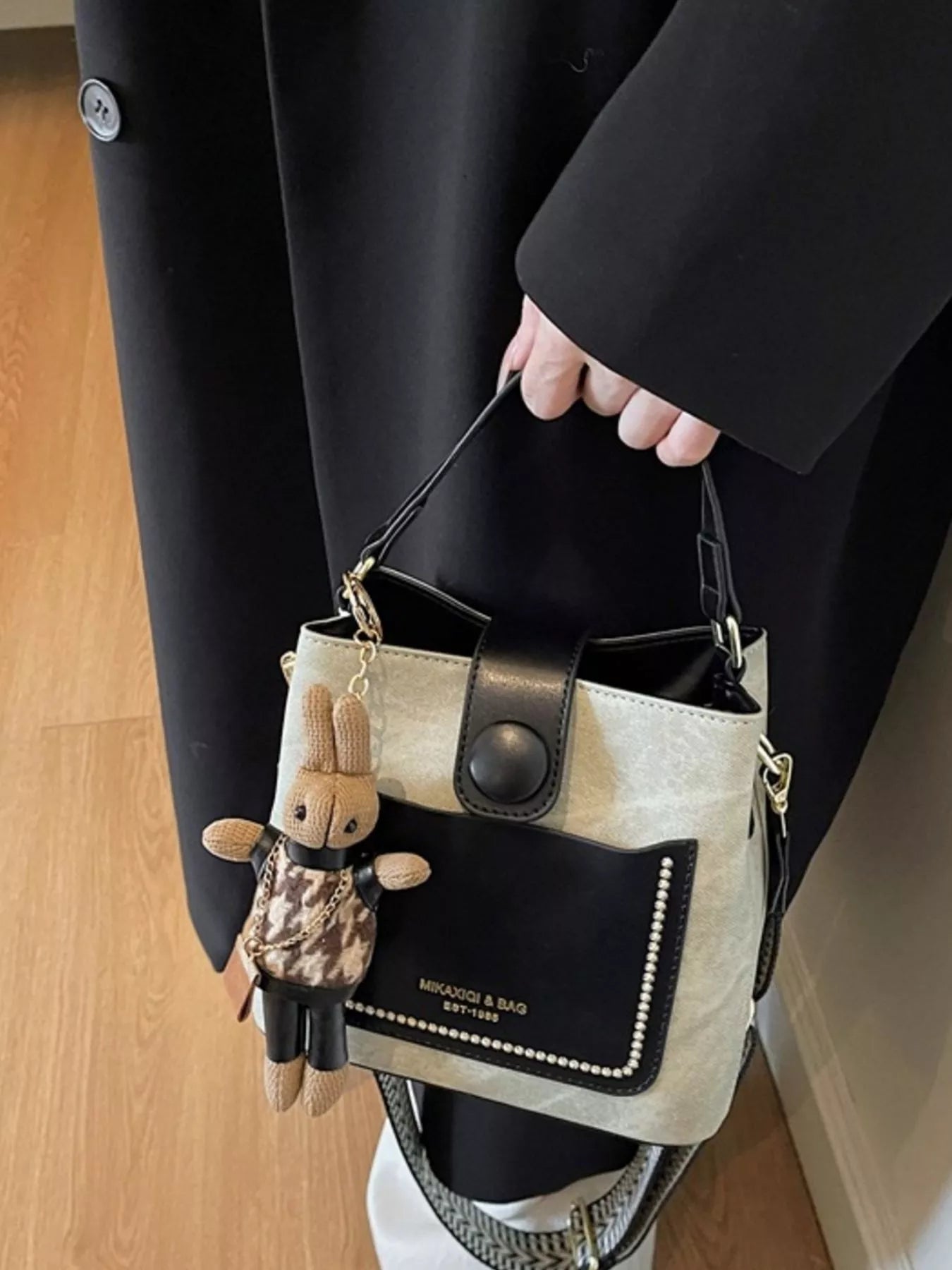 Person holding a black and beige handbag with a small plush toy attached, wearing a black coat.