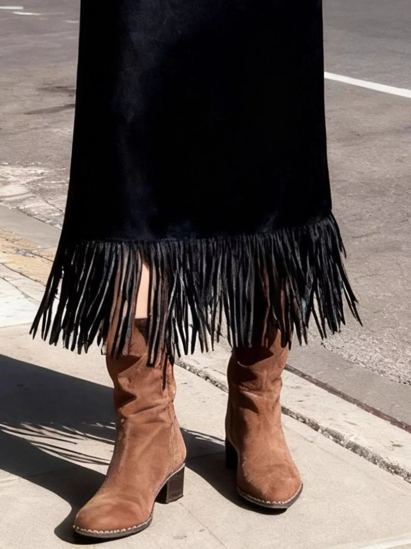 Brown suede boots with fringe on a black skirt against a street background