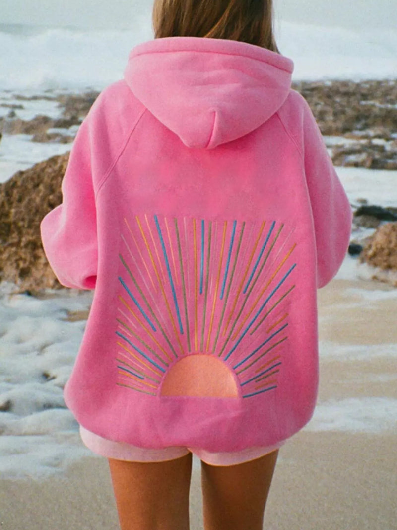 Person wearing a pink hoodie with a colorful design on the back, standing on a beach.