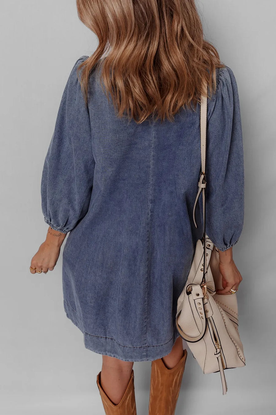 Person wearing a blue denim dress with brown boots and holding a beige handbag against a gray background