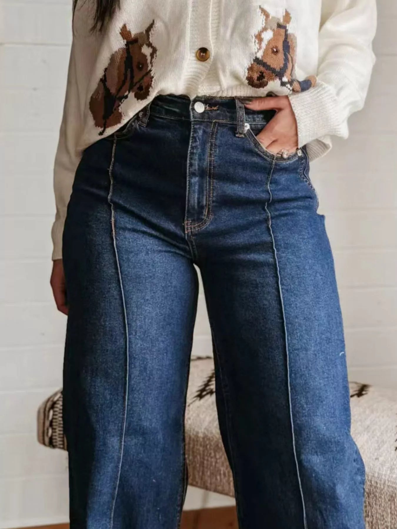 Person wearing a white sweater with horse designs and blue jeans indoors.
