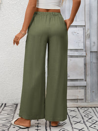 Person wearing olive green wide-leg pants with a white top and sandals, standing against a wooden panel background.