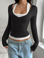 Person wearing a black long-sleeve top over a white tank top with blue jeans indoors.