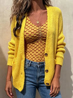 A woman wearing a bright yellow open-front knit cardigan over a mustard yellow patterned top, paired with blue jeans, standing against a light beige wall.