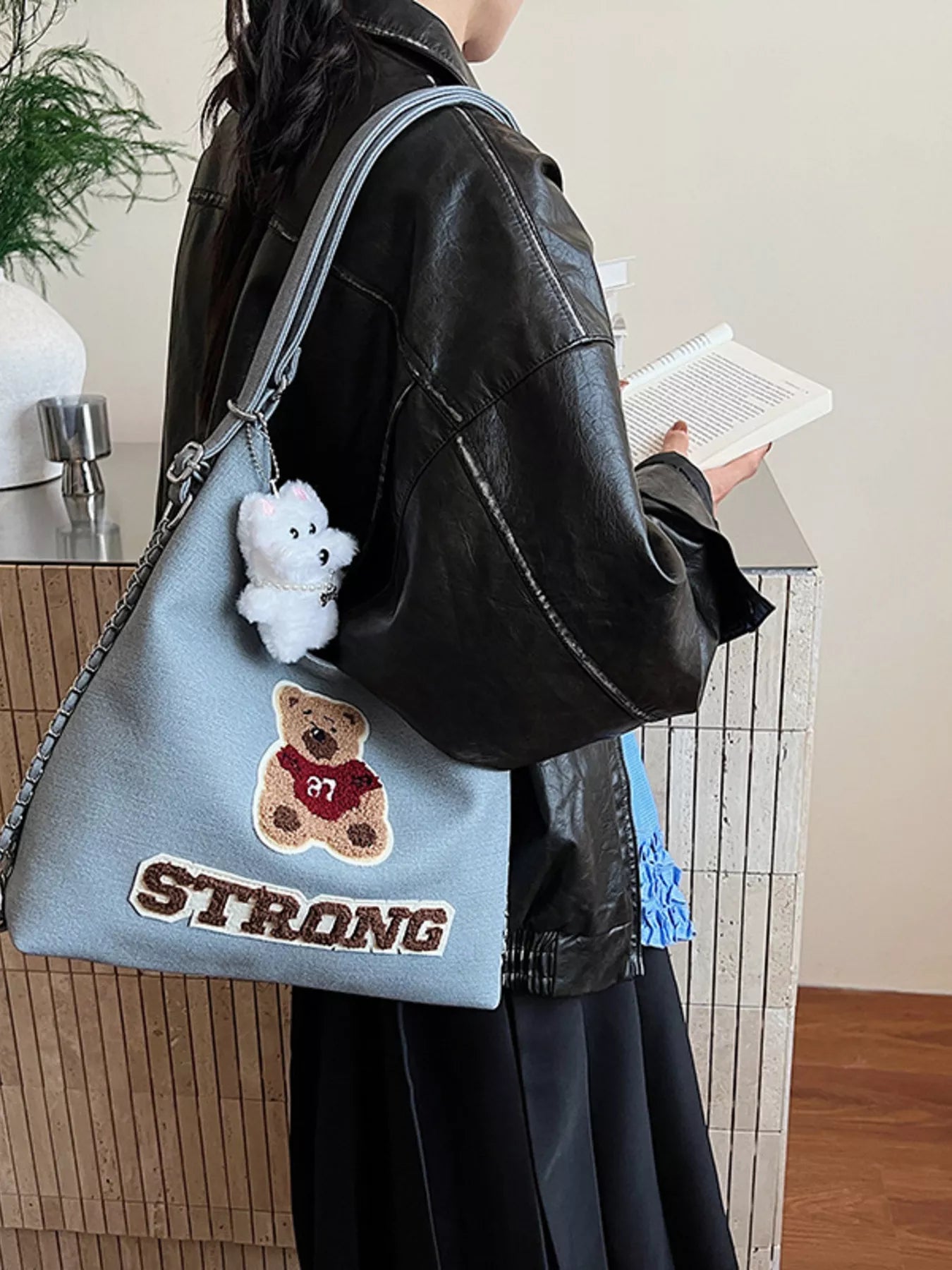 Person holding a blue bag with a teddy bear and 'STRONG' text, wearing a black leather jacket.