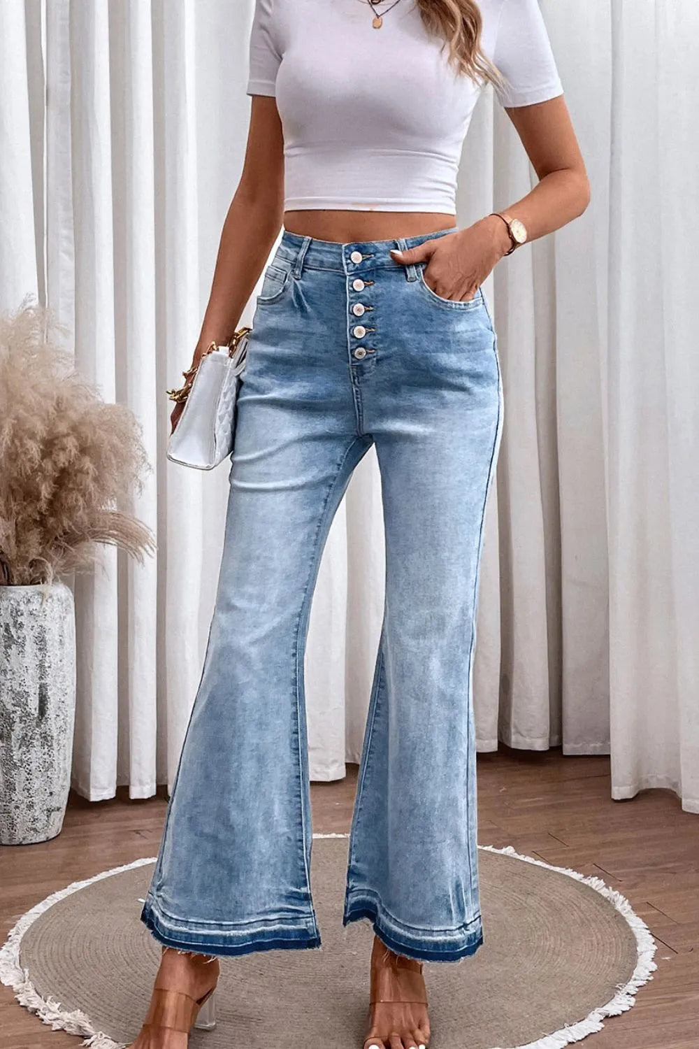 Woman wearing a white crop top and blue flared jeans in a room with curtains and a plant.