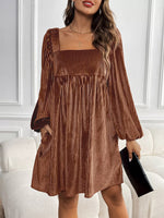 Woman wearing a brown pleated dress holding a black clutch against a white wall.