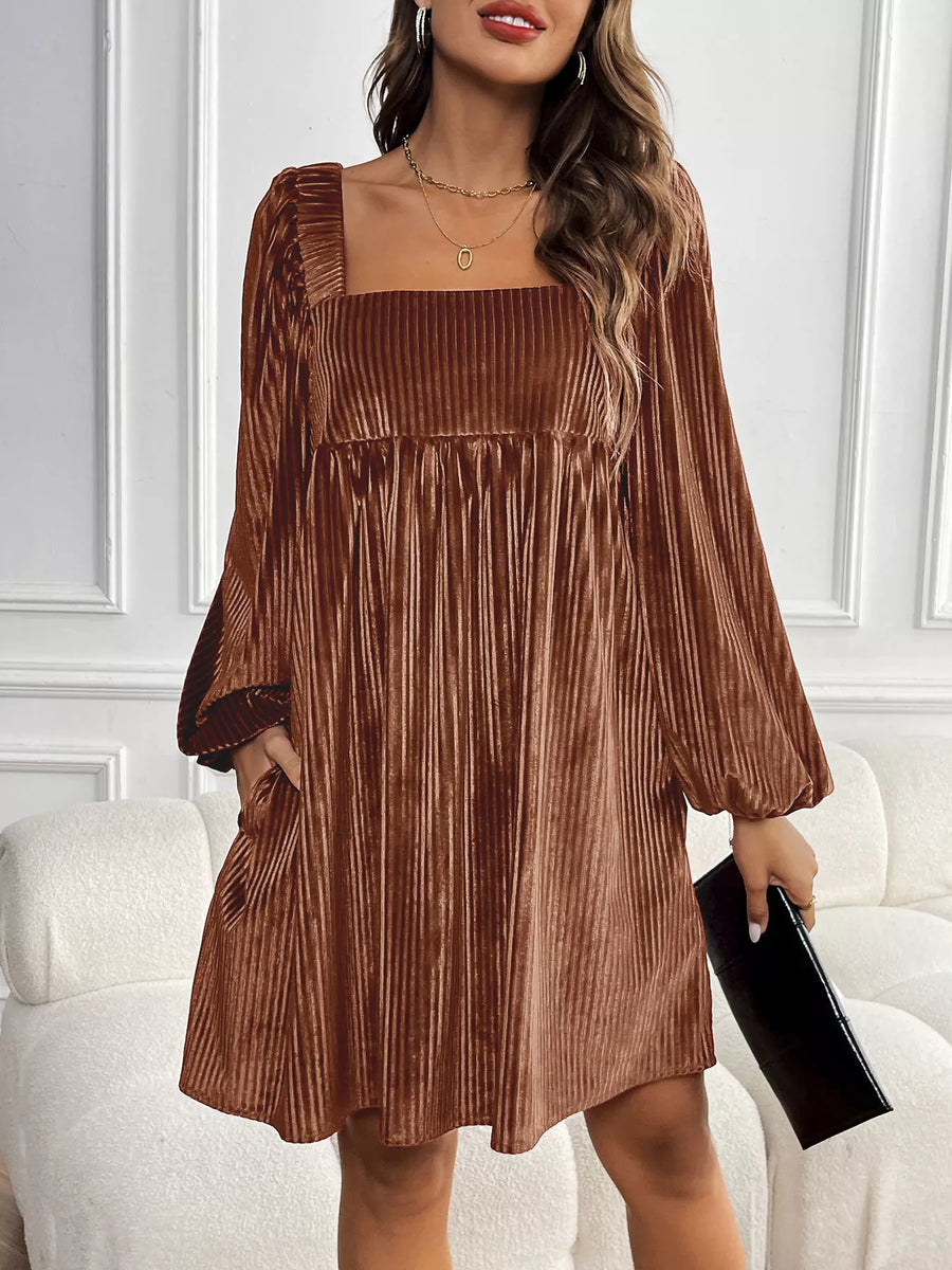 Woman wearing a brown pleated dress holding a black clutch against a white wall.