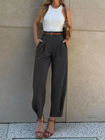 Woman wearing a white top, brown belt, and gray high-waisted pants against a neutral wall.