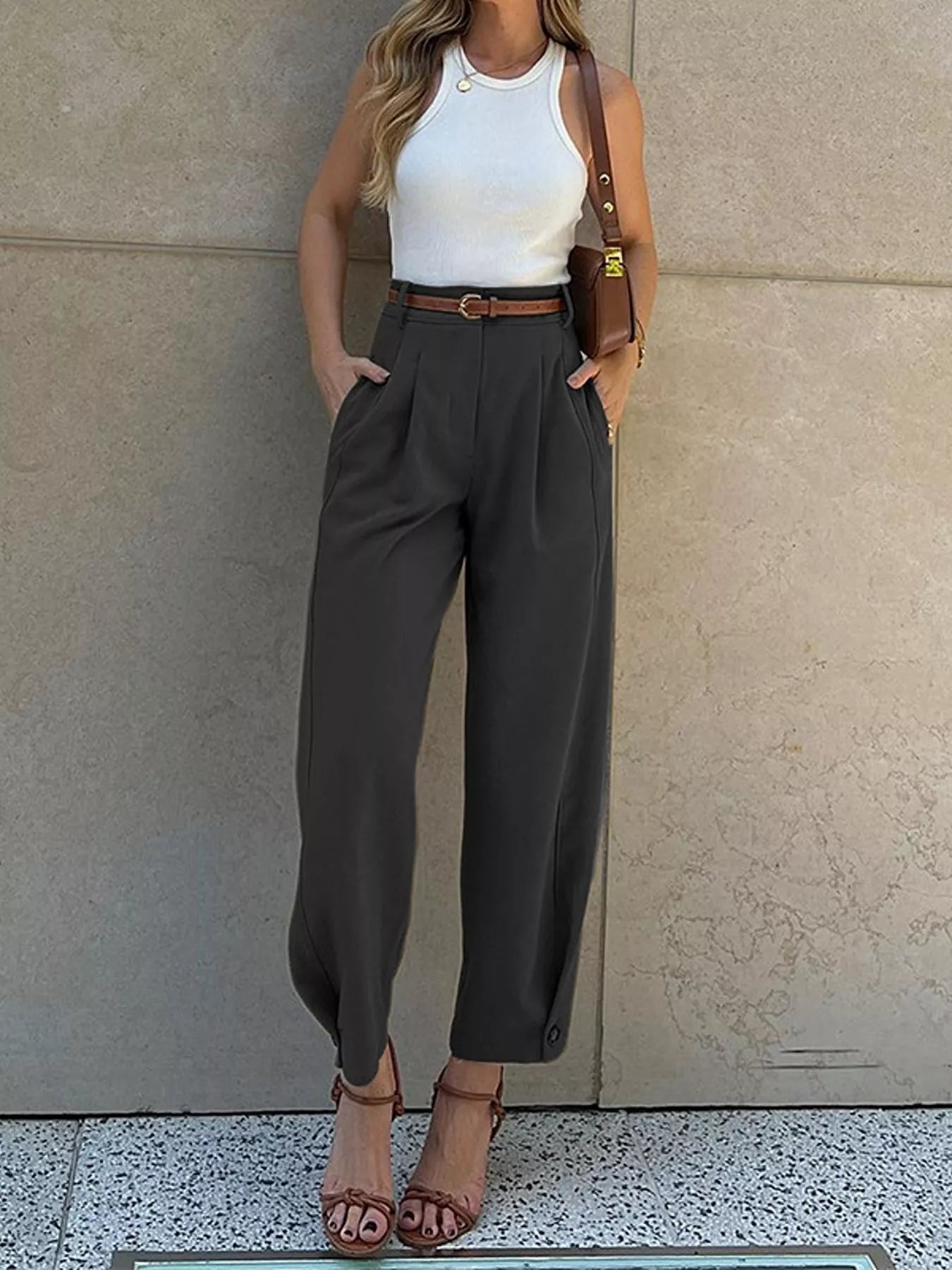 Woman wearing a white top, brown belt, and gray high-waisted pants against a neutral wall.
