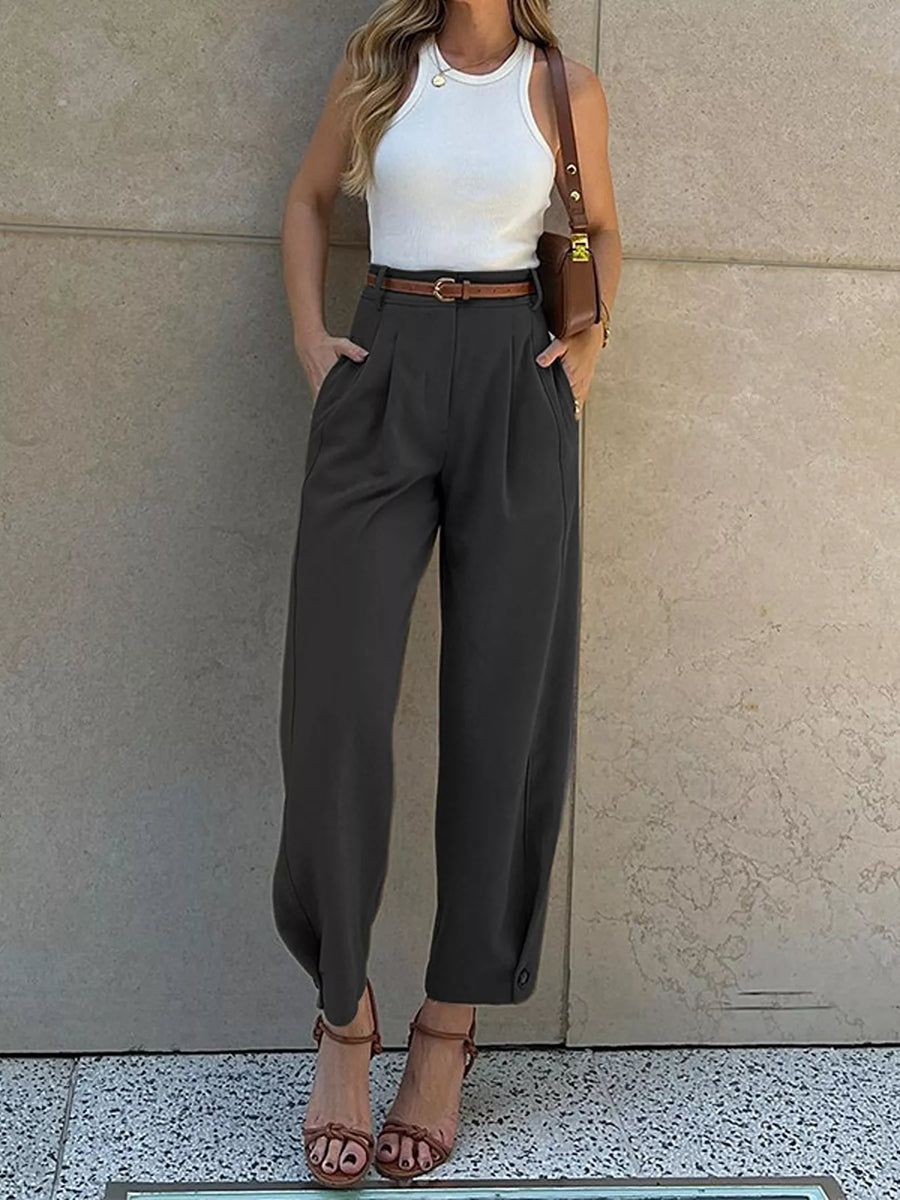 Woman wearing a white top, brown belt, and gray high-waisted pants against a neutral wall.