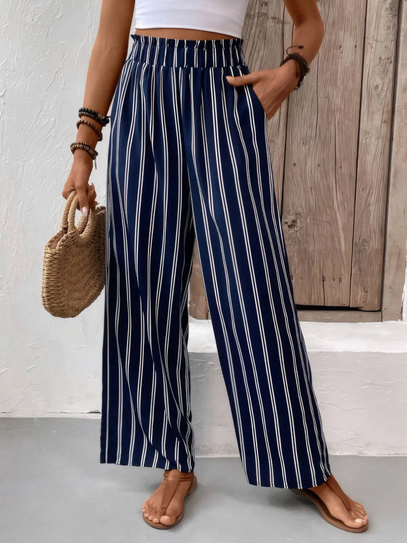 Person wearing navy and white striped pants with a white top and brown sandals, holding a woven bag.