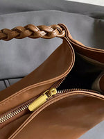 Brown leather handbag with braided handle and gold zipper on a gray background