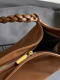 Brown leather handbag with braided handle and gold zipper on a gray background