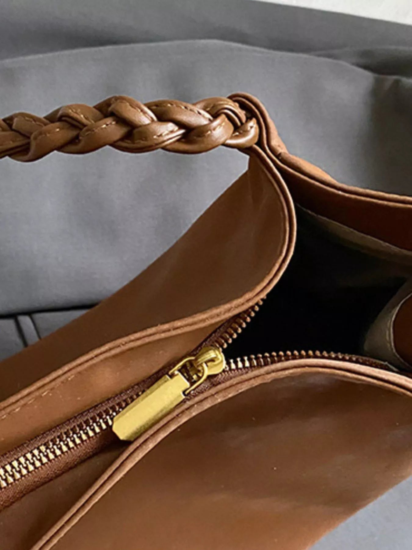 Brown leather handbag with braided handle and gold zipper on a gray background
