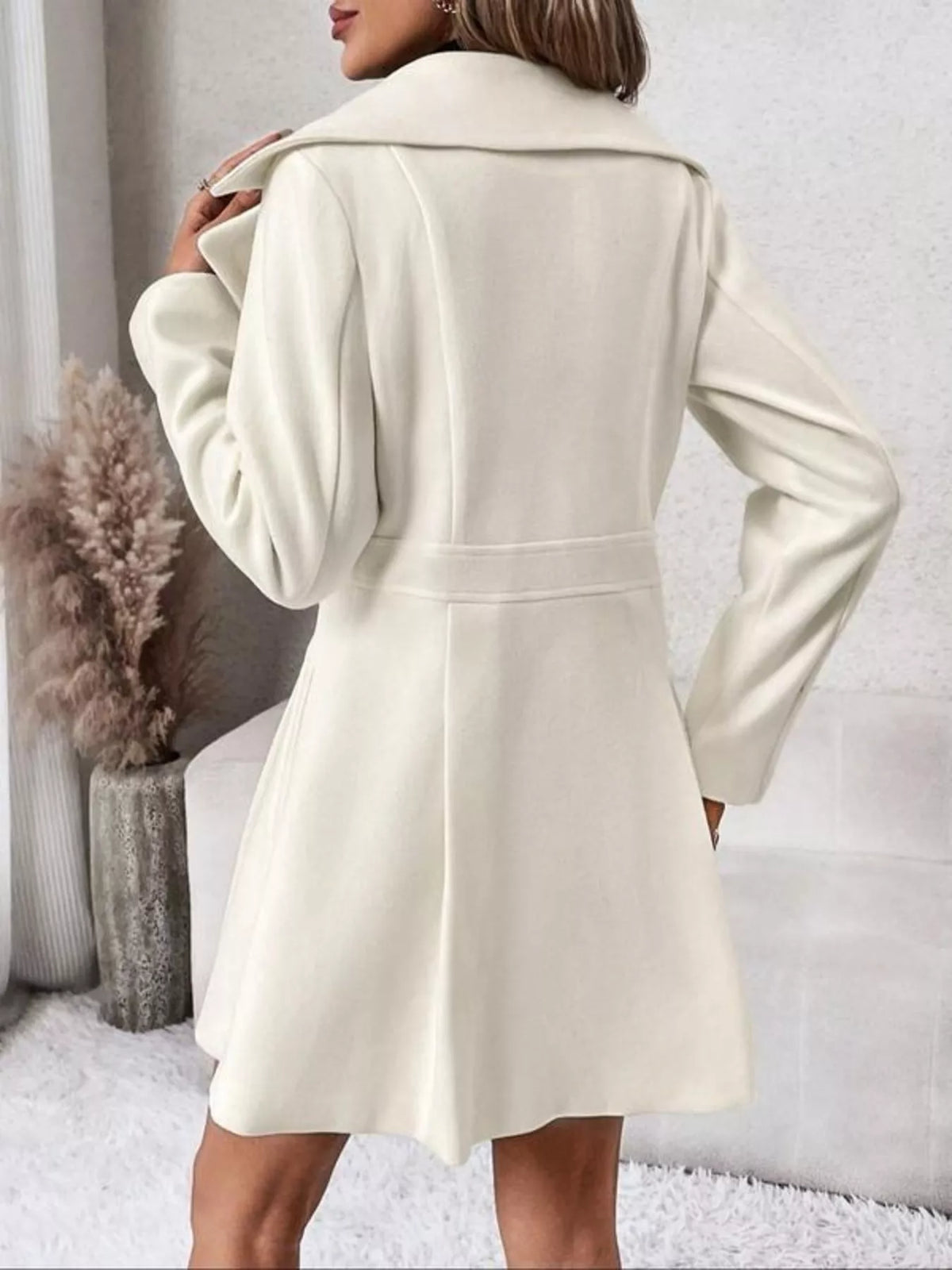 A woman wearing a cream-colored long coat with a wide collar and waist tie, shown from the back in an indoor setting.