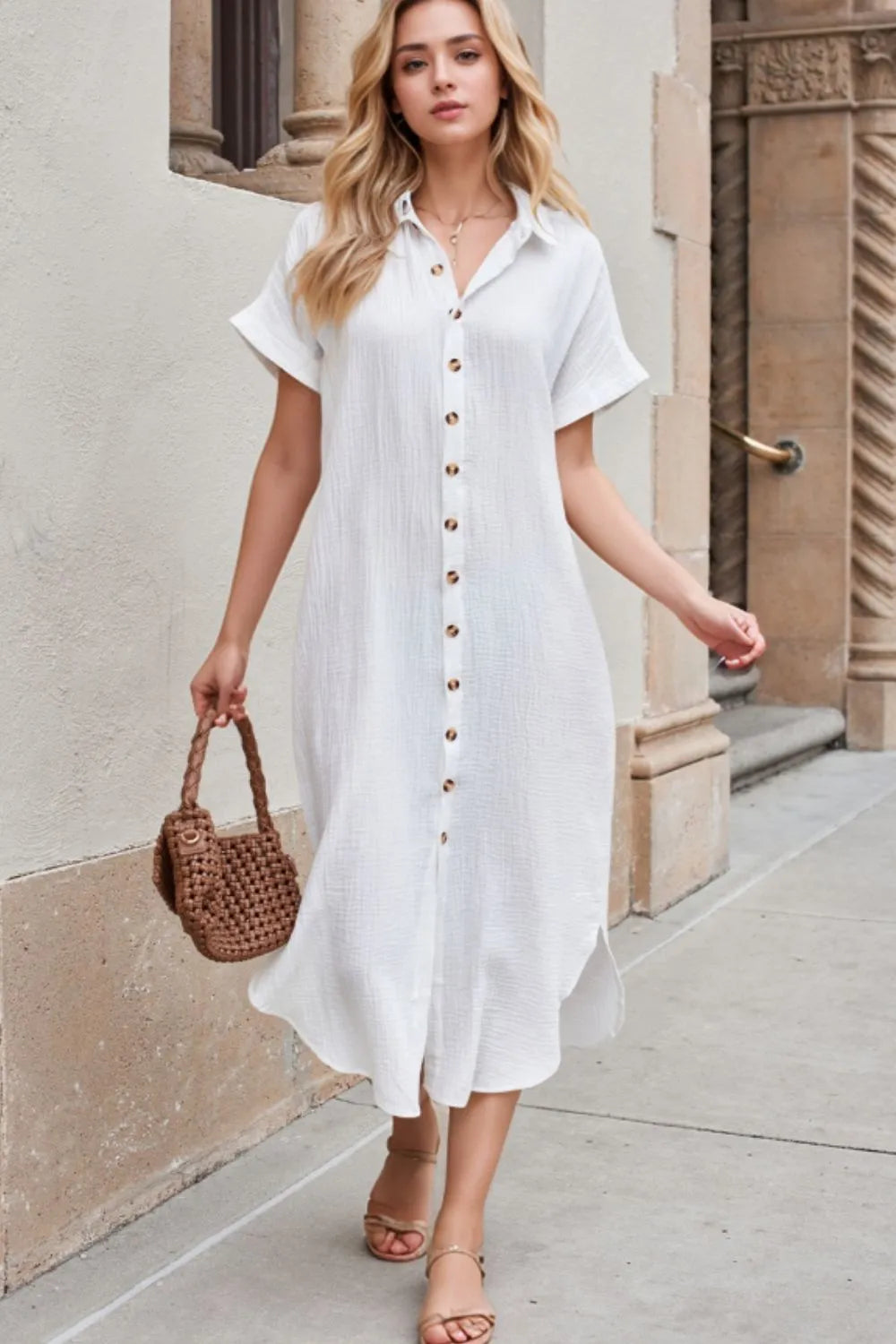 Woman wearing a white dress with buttons, holding a brown woven handbag, standing on a sidewalk.