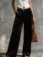 A model wearing black wide-leg velvet pants and a white cropped vest, holding a brown woven handbag against a textured gray wall.
