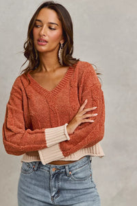 Woman wearing a rust-colored sweater with a beige trim and blue jeans on a gray background