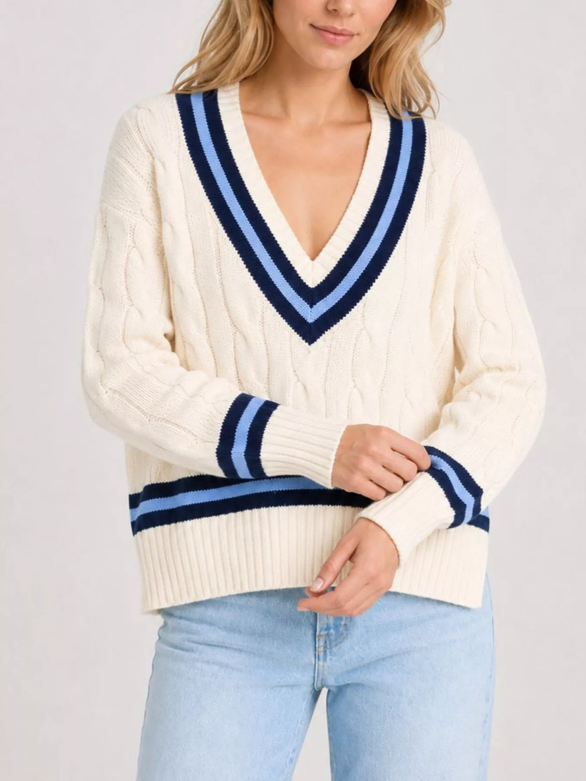 A woman wearing a cream cable-knit V-neck sweater with navy and light blue striped trim, paired with light blue jeans, against a light gray studio background.