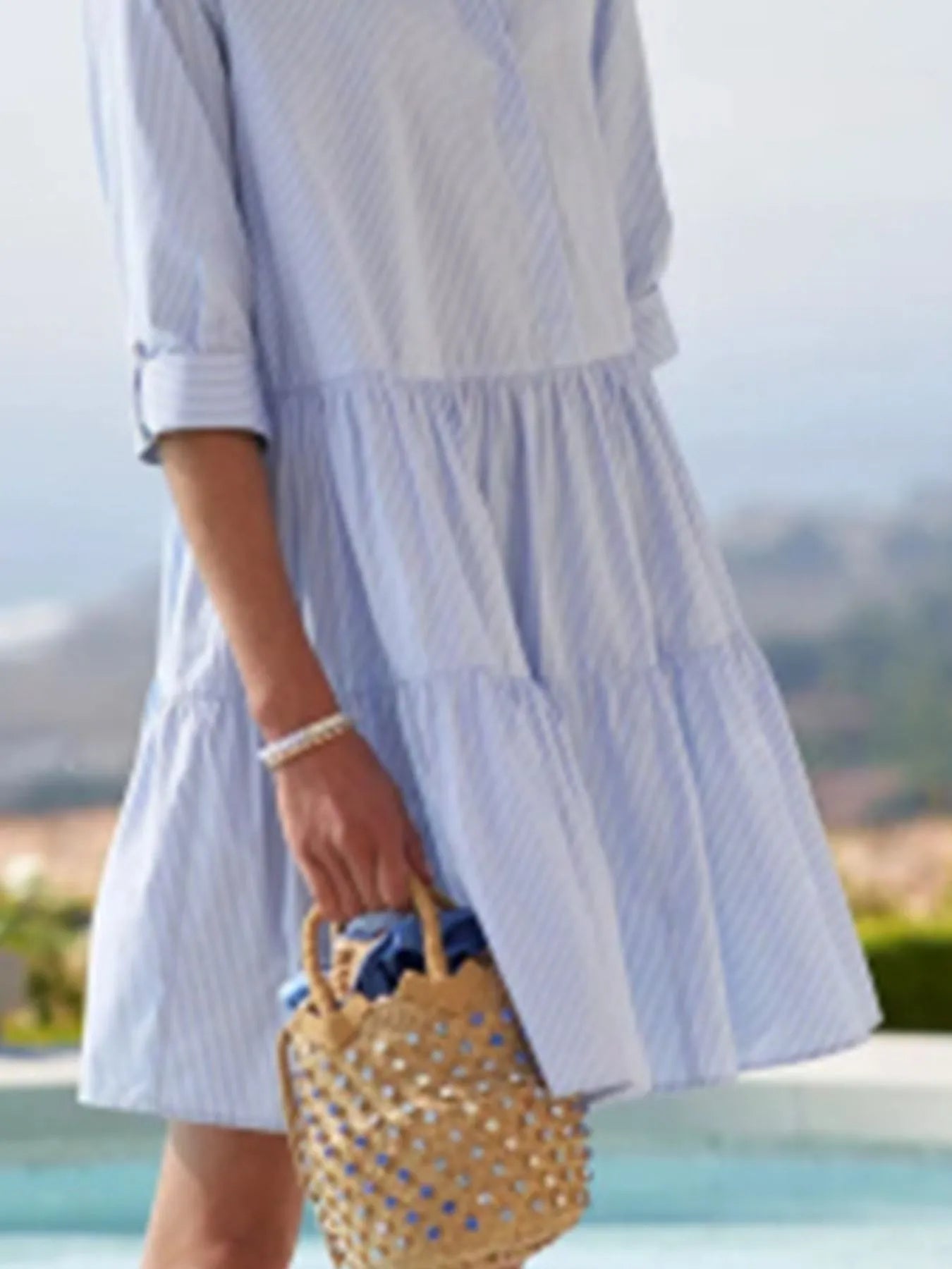 Person wearing a light blue dress holding a straw bag with a scenic background