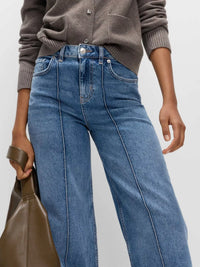 Person wearing blue jeans and a brown sweater with a brown leather bag on a light gray background
