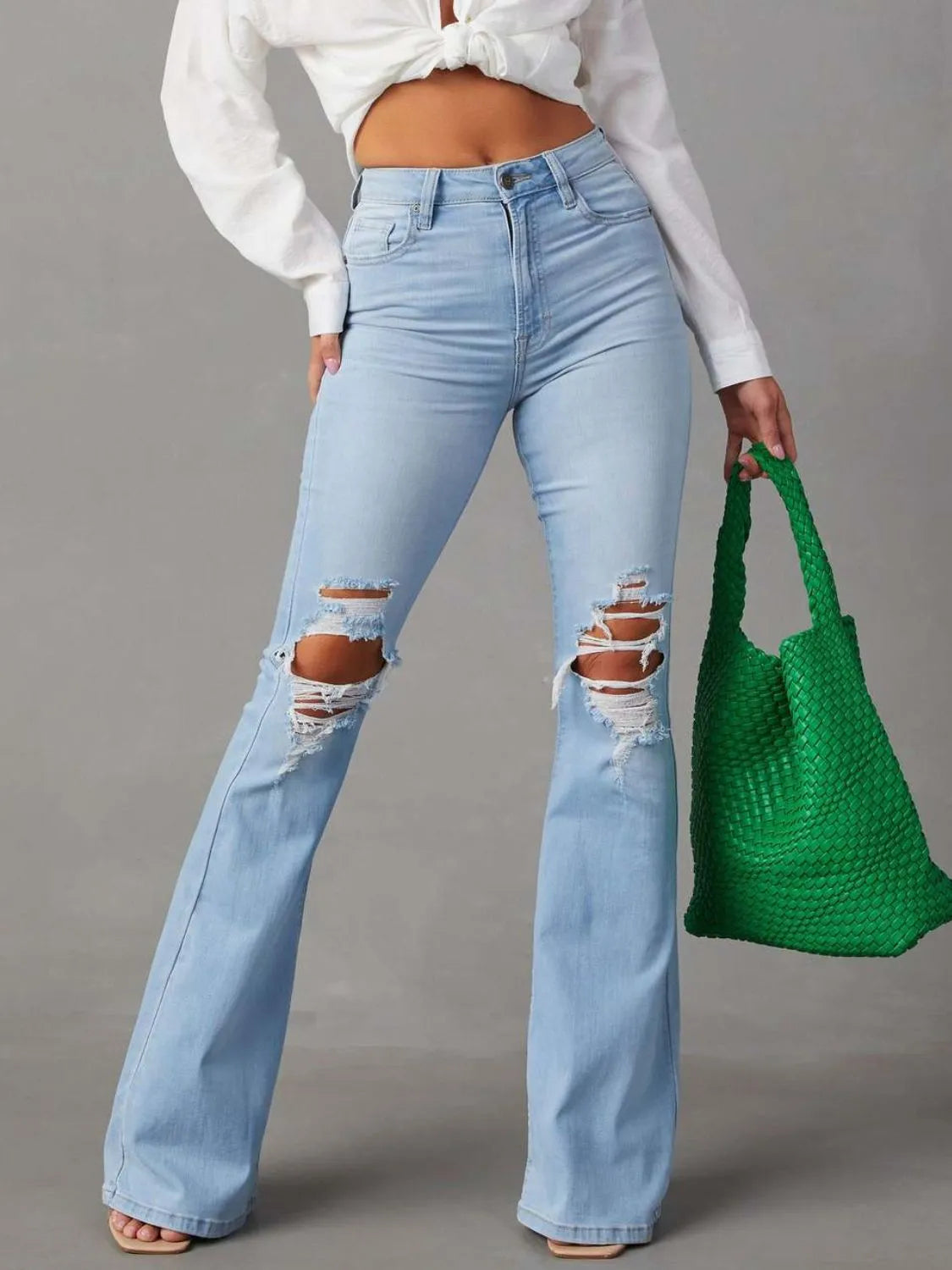 Person wearing light blue ripped jeans and a white cropped top, holding a green bag on a gray background