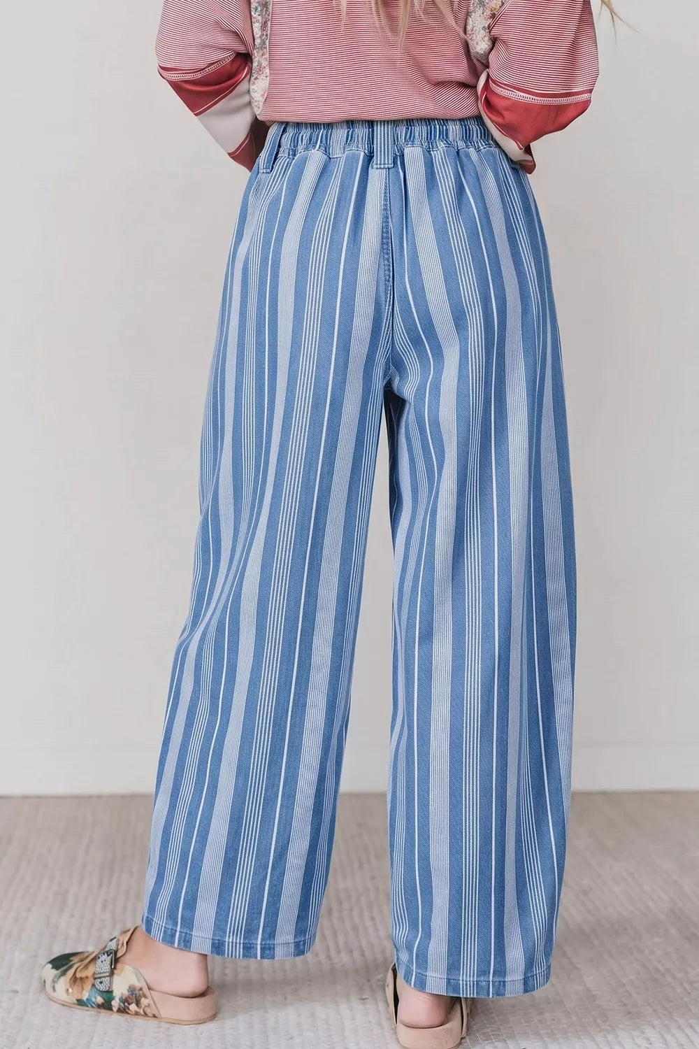 A person shown from the back wearing high-waisted, wide-leg blue and white striped trousers, a pink striped top, and floral mule sandals against a neutral indoor background.