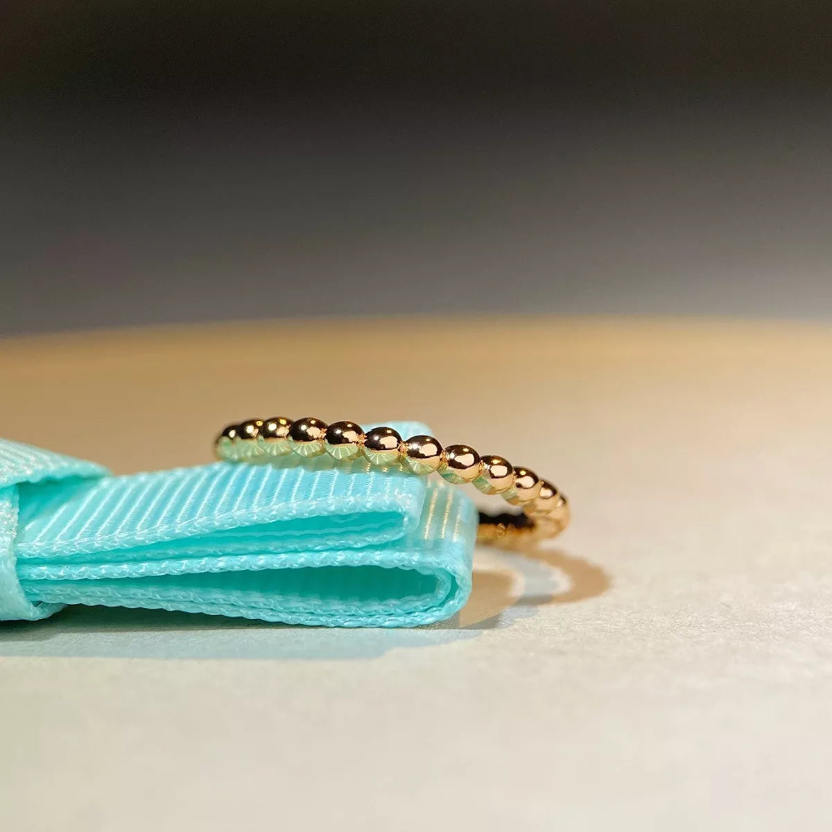 A delicate gold beaded ring resting on a light blue ribbon against a soft beige and gray background.