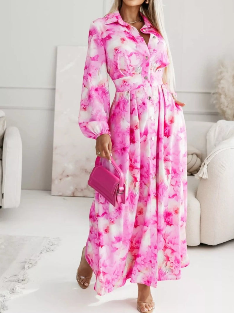 A woman wearing a long-sleeve pink and white floral maxi dress with a button-up front and waist tie, holding a matching pink handbag in a bright, modern living room setting.
