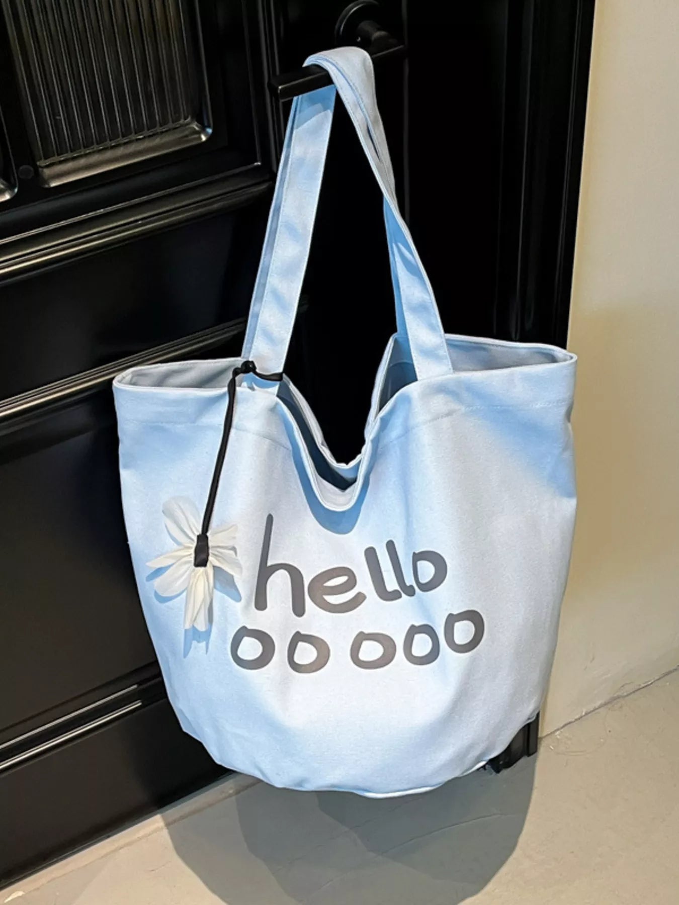 Light blue tote bag with 'hello ooo' text and flower design on a door step.