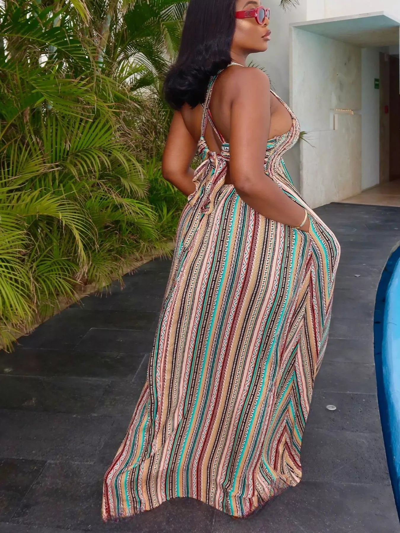 Woman in a colorful striped dress standing outdoors with greenery and a building in the background.