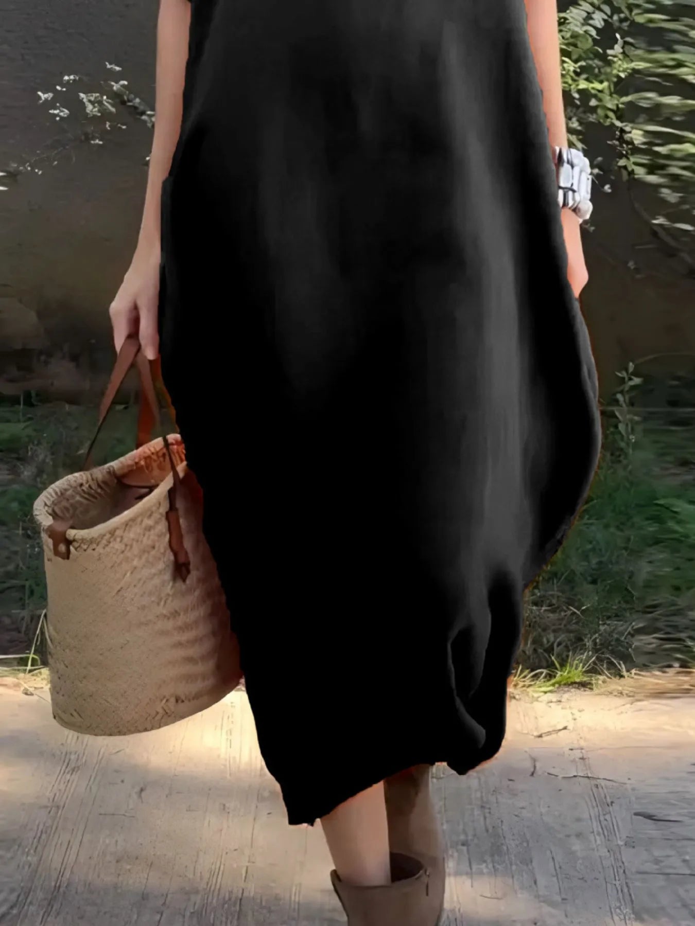 Person wearing a black dress holding a woven bag outdoors.