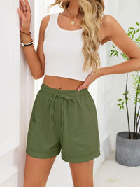 Woman wearing a white crop top and green shorts against a white wall with a plant.