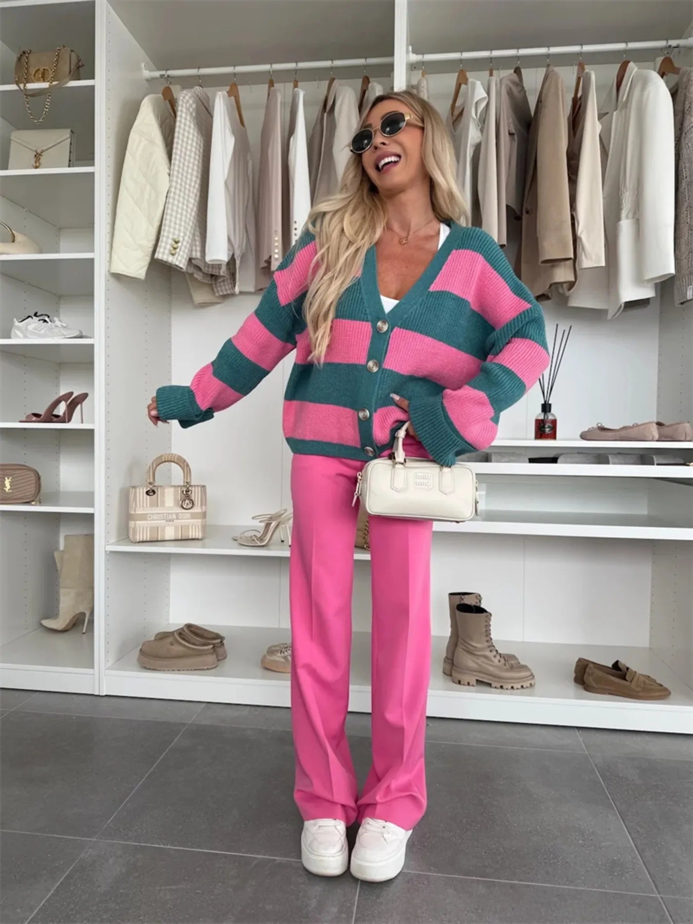 Woman in a pink and green striped cardigan and pink pants standing in a wardrobe with clothes and shoes on shelves.