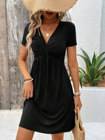 Woman wearing a black dress with a straw hat and bag, standing against a wooden wall.
