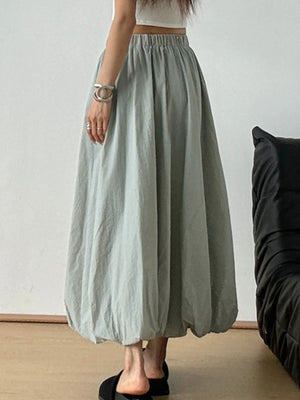 Person wearing a long light gray skirt with a white top and black shoes on a neutral background.