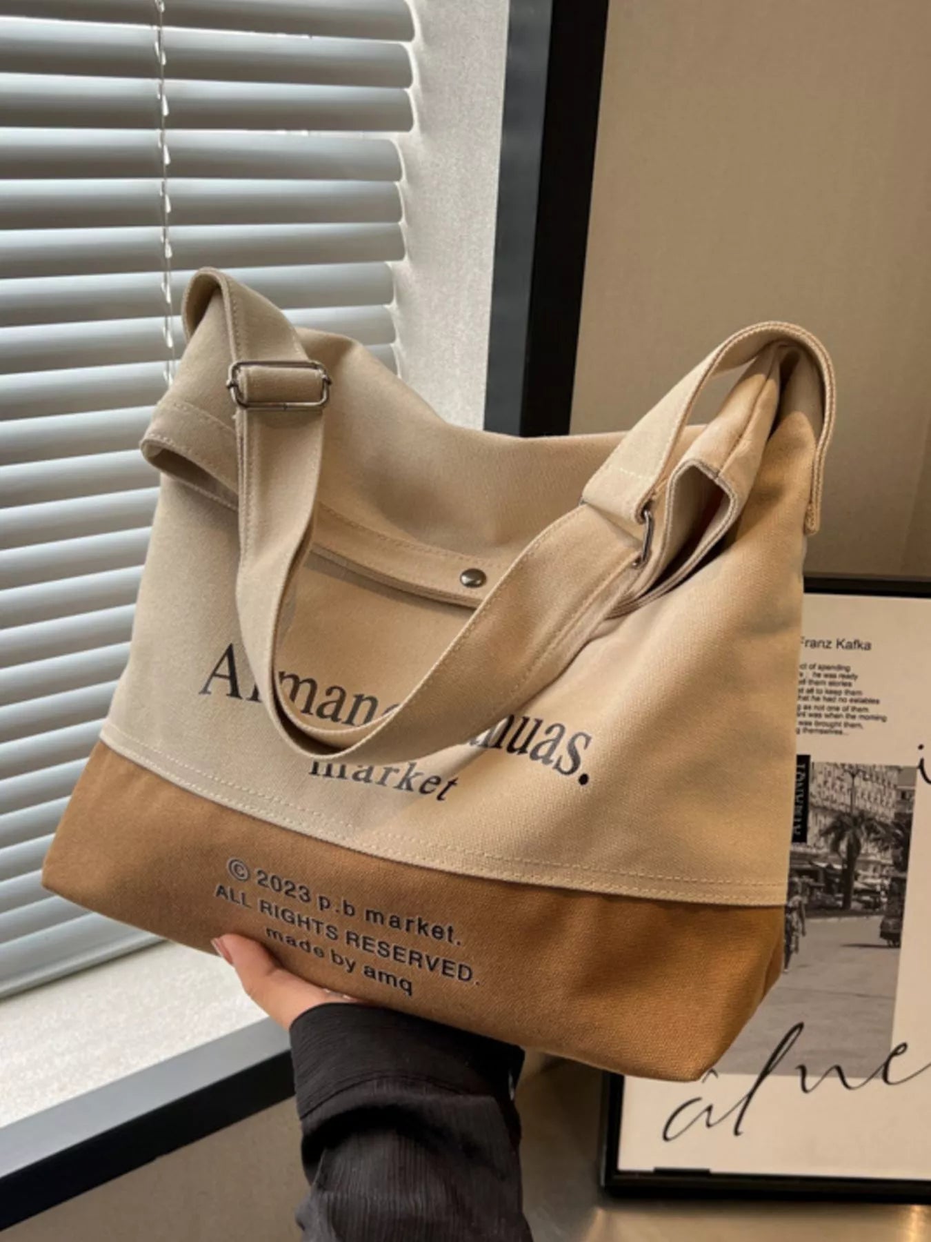 Beige and brown tote bag held by a person with visible branding, against a neutral background.