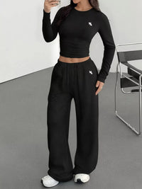 Person wearing a black long-sleeve crop top and wide-leg pants with a small logo, standing in a minimalistic room.