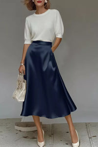 Woman wearing a white top and navy blue skirt holding a white handbag against a gray background