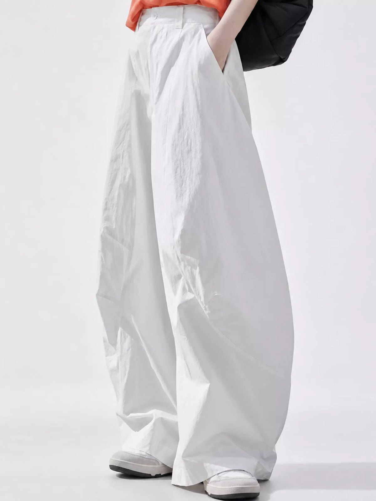 A person wearing very wide-leg white pants with a relaxed, baggy fit, paired with a coral-orange top and white sneakers, shown from the waist down against a light gray studio background.