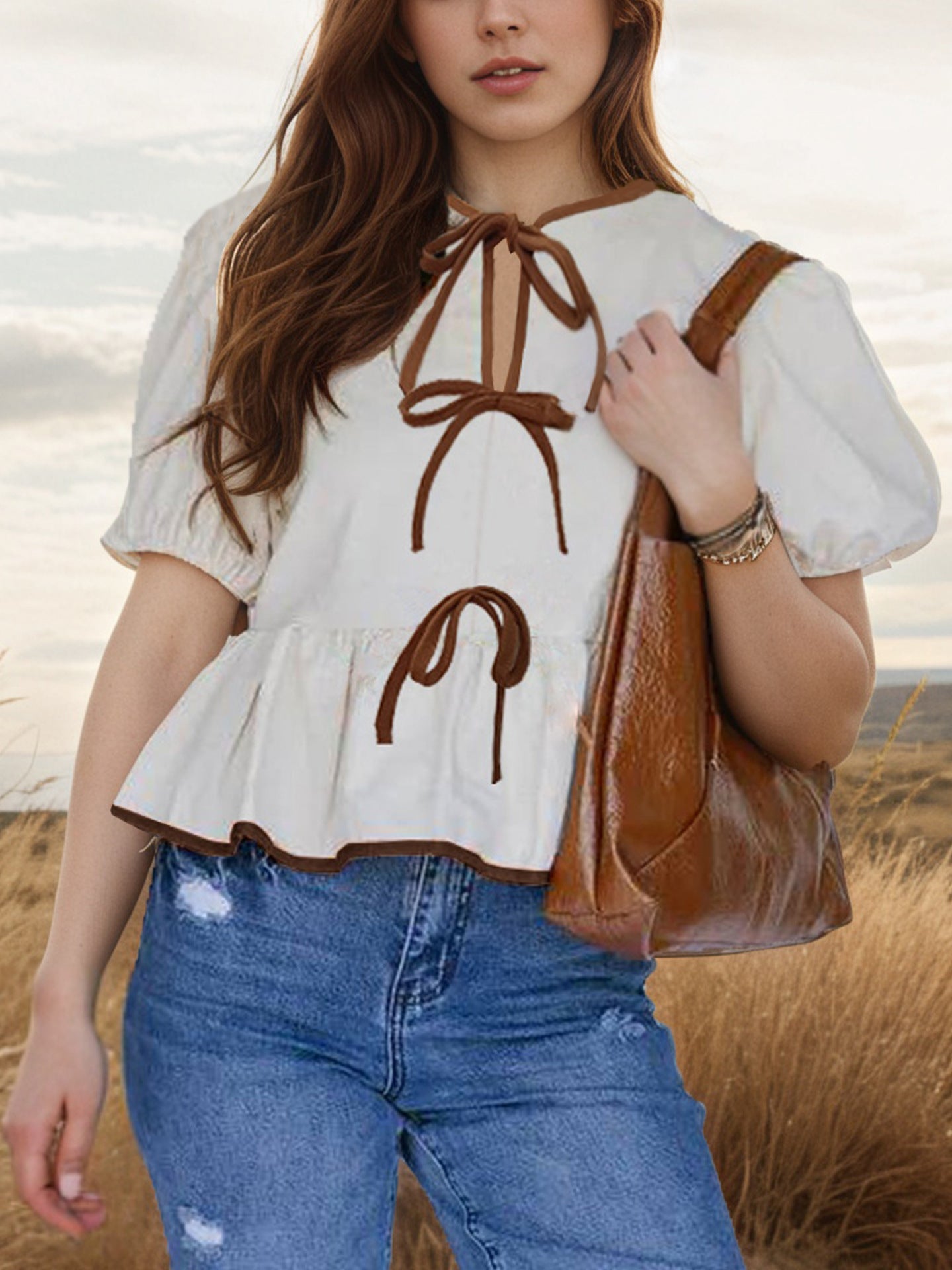 Woman wearing a white blouse with brown embroidery and blue jeans, holding a brown handbag in a field.