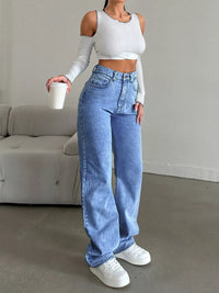 Person wearing a white long-sleeve crop top and blue jeans, holding a white cup in a minimalistic room.