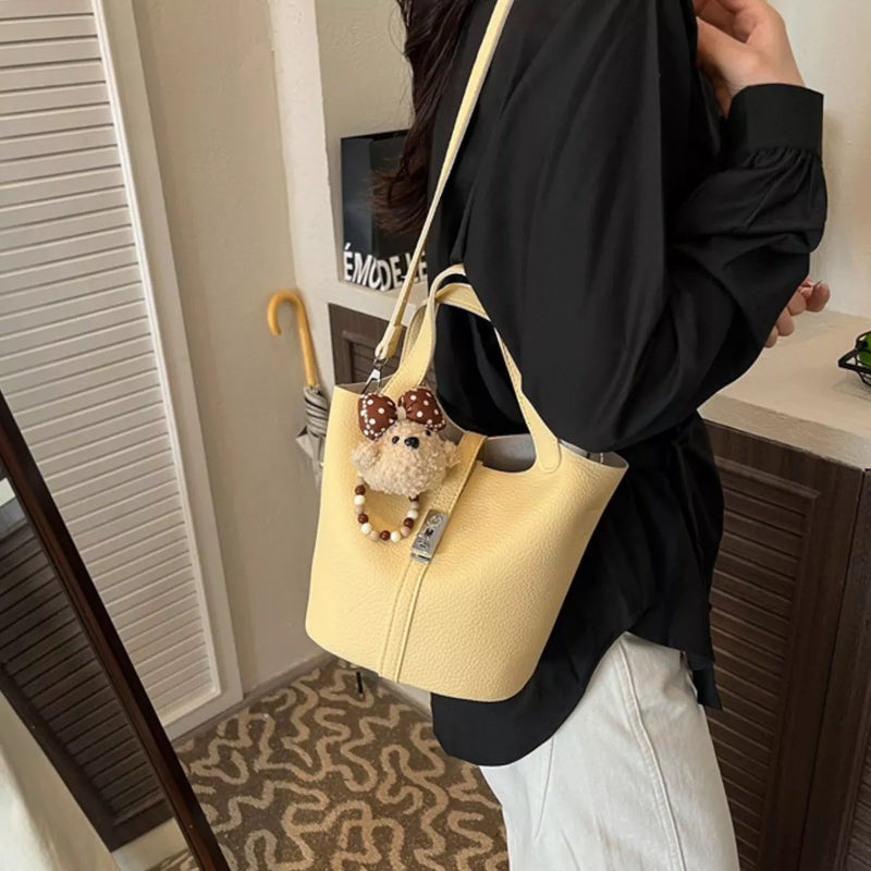 A person wearing a black blouse and white pants carries a light yellow pebbled leather bucket bag with a shoulder strap. The bag features a small plush bear charm with a brown polka-dot bow and a beaded wristlet loop.